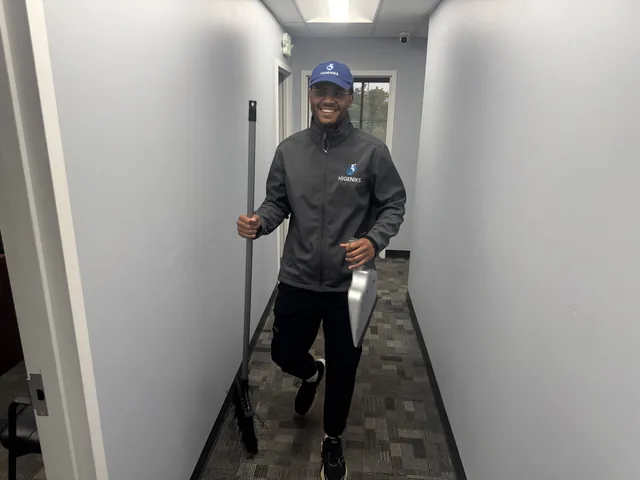 A Higeniks cleaner in branded uniform carrying a broom along an office hallway.